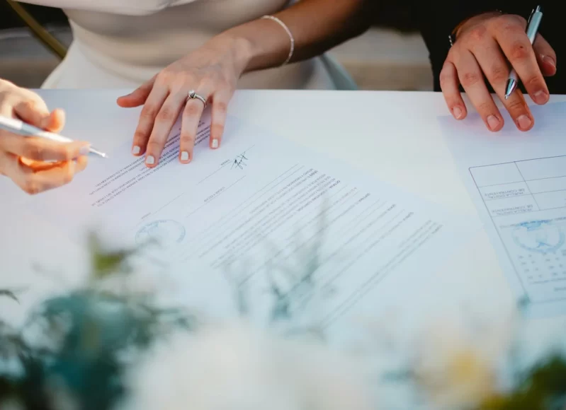 Imagem de um casal assinando documentos no casamento civil, em uma mesa simples, com caneta em mãos e clima formal.