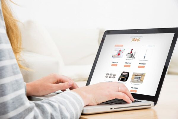 Pessoa navegando em notebook por uma lista online de presentes de casamento, com itens domésticos e valores exibidos na tela.