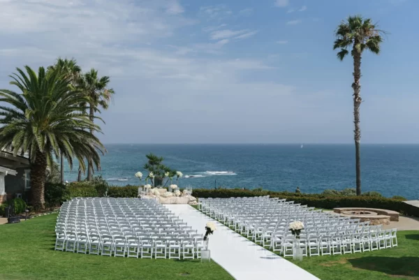 Cerimônia de casamento ao ar livre com cadeiras brancas, decoração floral e vista para o mar.
