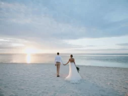 Casal de noivos caminhando de mãos dadas pela praia ao pôr do sol.