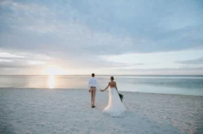 Casal de noivos caminhando de mãos dadas pela praia ao pôr do sol.