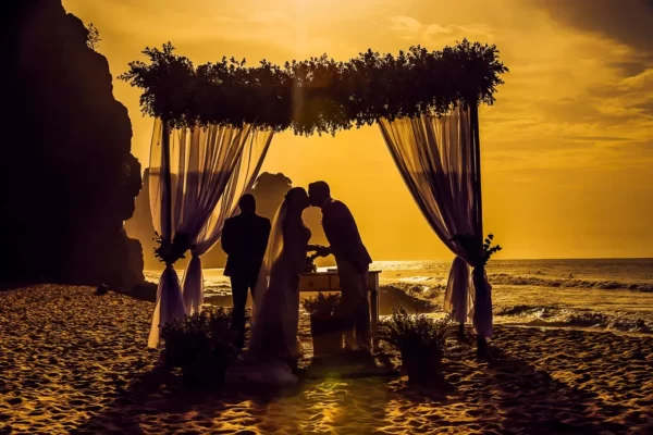 Casal de noivos se beijando durante cerimônia de casamento na praia ao pôr do sol.