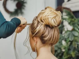 Imagem mostra uma mulher em um salão de beleza, fazendo um penteado com a ajuda de um profissional, que trabalha cuidadosamente em seu cabelo