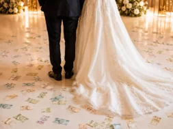 Noivos vistos de costas em festa de casamento, com notas de dinheiro espalhadas pelo chão ao redor do vestido da noiva.