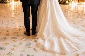 Noivos vistos de costas em festa de casamento, com notas de dinheiro espalhadas pelo chão ao redor do vestido da noiva.