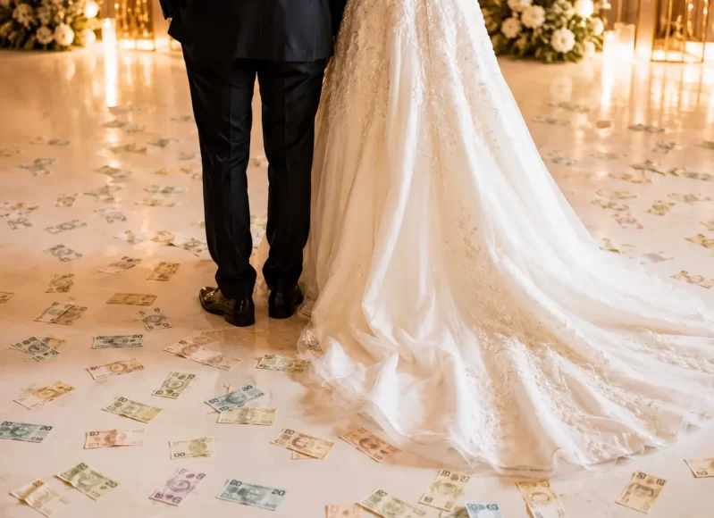 Noivos vistos de costas em festa de casamento, com notas de dinheiro espalhadas pelo chão ao redor do vestido da noiva.