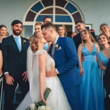 Noivos se beijam na saída da cerimônia, cercados por padrinhos e madrinhas com trajes em tons de azul.