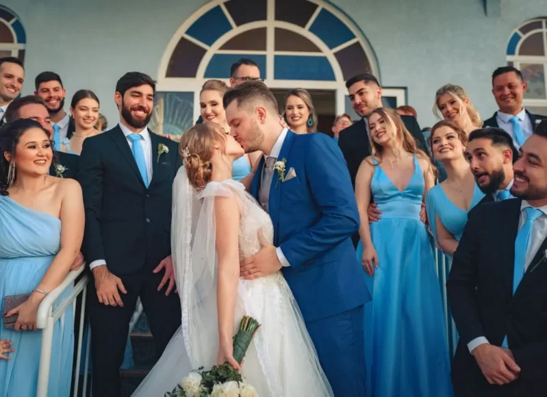 Noivos se beijam na saída da cerimônia, cercados por padrinhos e madrinhas com trajes em tons de azul.