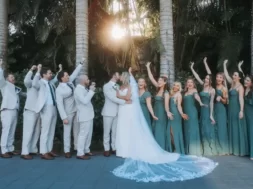 Imagem de um casal de noivos no centro, cercado por vários padrinhos e madrinhas, com árvores ao fundo.