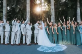 Imagem de um casal de noivos no centro, cercado por vários padrinhos e madrinhas, com árvores ao fundo.