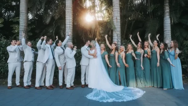 Imagem de um casal de noivos no centro, cercado por vários padrinhos e madrinhas, com árvores ao fundo.