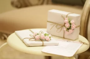 Imagem de dois presentes brancos decorados com flores rosas e barbante sobre uma mesa, com um envelope de carta.