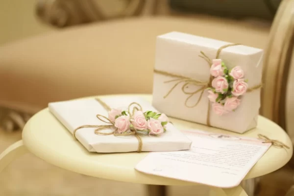 Imagem de dois presentes brancos decorados com flores rosas e barbante sobre uma mesa, com um envelope de carta.