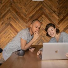 Casal sentado à mesa usando notebook e celular, sorrindo enquanto monta uma lista de casamento divertida em ambiente aconchegante.