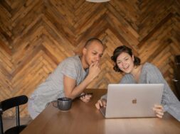 Casal sentado à mesa usando notebook e celular, sorrindo enquanto monta uma lista de casamento divertida em ambiente aconchegante.