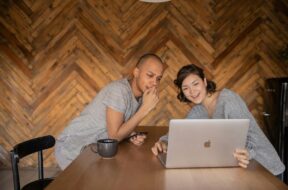 Casal sentado à mesa usando notebook e celular, sorrindo enquanto monta uma lista de casamento divertida em ambiente aconchegante.