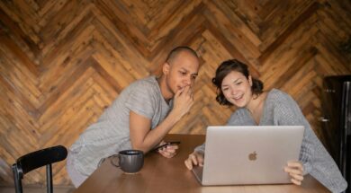 Casal sentado à mesa usando notebook e celular, sorrindo enquanto monta uma lista de casamento divertida em ambiente aconchegante.