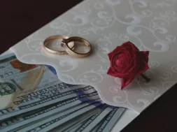 Imagem de um convite de casamento com dinheiro dentro, sobre o convite há alianças de casamento e uma rosa.