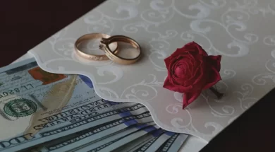 Imagem de um convite de casamento com dinheiro dentro, sobre o convite há alianças de casamento e uma rosa.