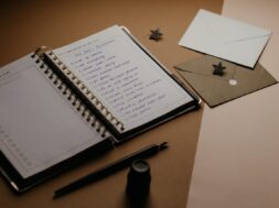 Imagem de um caderno aberto com uma lista para chá de cozinha, ao lado há envelopes de convite e canetas sobre a mesa.