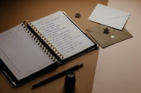 Imagem de um caderno aberto com uma lista para chá de cozinha, ao lado há envelopes de convite e canetas sobre a mesa.