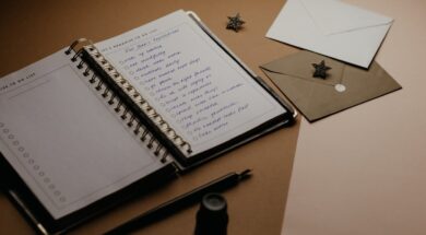 Imagem de um caderno aberto com uma lista para chá de cozinha, ao lado há envelopes de convite e canetas sobre a mesa.