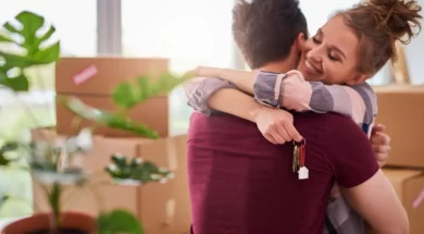 Imagem de um casal se abraçando, com caixas de mudança ao fundo, aparentando ser um casal feliz com a casa nova.