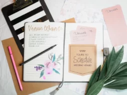 Imagem de um caderno aberto com checklist para casamento, com caneta em cima, cartões escritos, folhas verdes decorativas ao lado e itens de papelaria sobre a mesa.