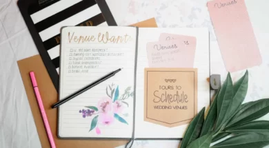Imagem de um caderno aberto com checklist para casamento, com caneta em cima, cartões escritos, folhas verdes decorativas ao lado e itens de papelaria sobre a mesa.