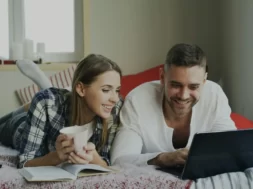 Imagem de um casal sobre a cama mexendo no notebook.