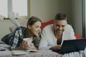 Imagem de um casal sobre a cama mexendo no notebook.