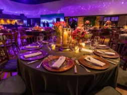 Imagem mostra mesa de evento elegante, com flores, velas e iluminação roxa em ambiente sofisticado.