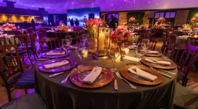 Imagem mostra mesa de evento elegante, com flores, velas e iluminação roxa em ambiente sofisticado.
