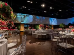 A imagem mostra um salão de casamento elegante, com mesas decoradas, flores em tons de vermelho e um grande painel ao fundo com fotos do casal.