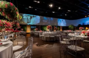 A imagem mostra um salão de casamento elegante, com mesas decoradas, flores em tons de vermelho e um grande painel ao fundo com fotos do casal.