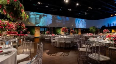 A imagem mostra um salão de casamento elegante, com mesas decoradas, flores em tons de vermelho e um grande painel ao fundo com fotos do casal.