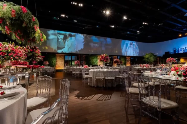 A imagem mostra um salão de casamento elegante, com mesas decoradas, flores em tons de vermelho e um grande painel ao fundo com fotos do casal.