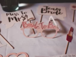 Óculos decorativos de despedida de solteira com a frase “Bride to Be”, cercados por plaquinhas temáticas e um acessório em formato de batom.