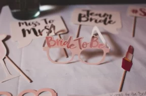Óculos decorativos de despedida de solteira com a frase “Bride to Be”, cercados por plaquinhas temáticas e um acessório em formato de batom.