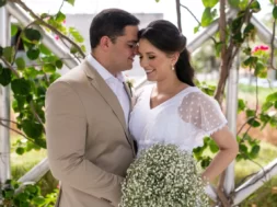 Casal de noivos sorrindo durante cerimônia de casamento ao ar livre, com buquê de mosquitinho e decoração com folhagens.