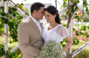 Casal de noivos sorrindo durante cerimônia de casamento ao ar livre, com buquê de mosquitinho e decoração com folhagens.