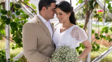 Casal de noivos sorrindo durante cerimônia de casamento ao ar livre, com buquê de mosquitinho e decoração com folhagens.