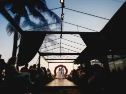 Casal durante cerimônia de casamento ao ar livre, diante de um arco de flores e cercado por convidados ao entardecer.