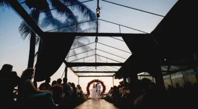 Casal durante cerimônia de casamento ao ar livre, diante de um arco de flores e cercado por convidados ao entardecer.