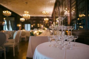 Taças de champanhe empilhadas sobre uma mesa em um elegante salão de festas decorado para casamento ou evento, com mesas, cadeiras e lustres ao fundo.