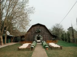 Cerimônia de casamento rústica ao ar livre, com bancos de madeira, flores brancas e celeiro ao fundo.