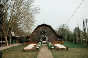 Cerimônia de casamento rústica ao ar livre, com bancos de madeira, flores brancas e celeiro ao fundo.