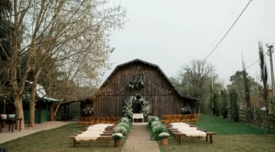 Cerimônia de casamento rústica ao ar livre, com bancos de madeira, flores brancas e celeiro ao fundo.