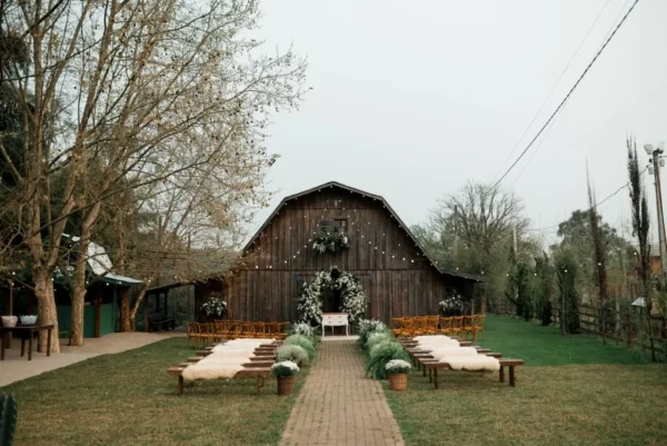 Cerimônia de casamento rústica ao ar livre, com bancos de madeira, flores brancas e celeiro ao fundo.
